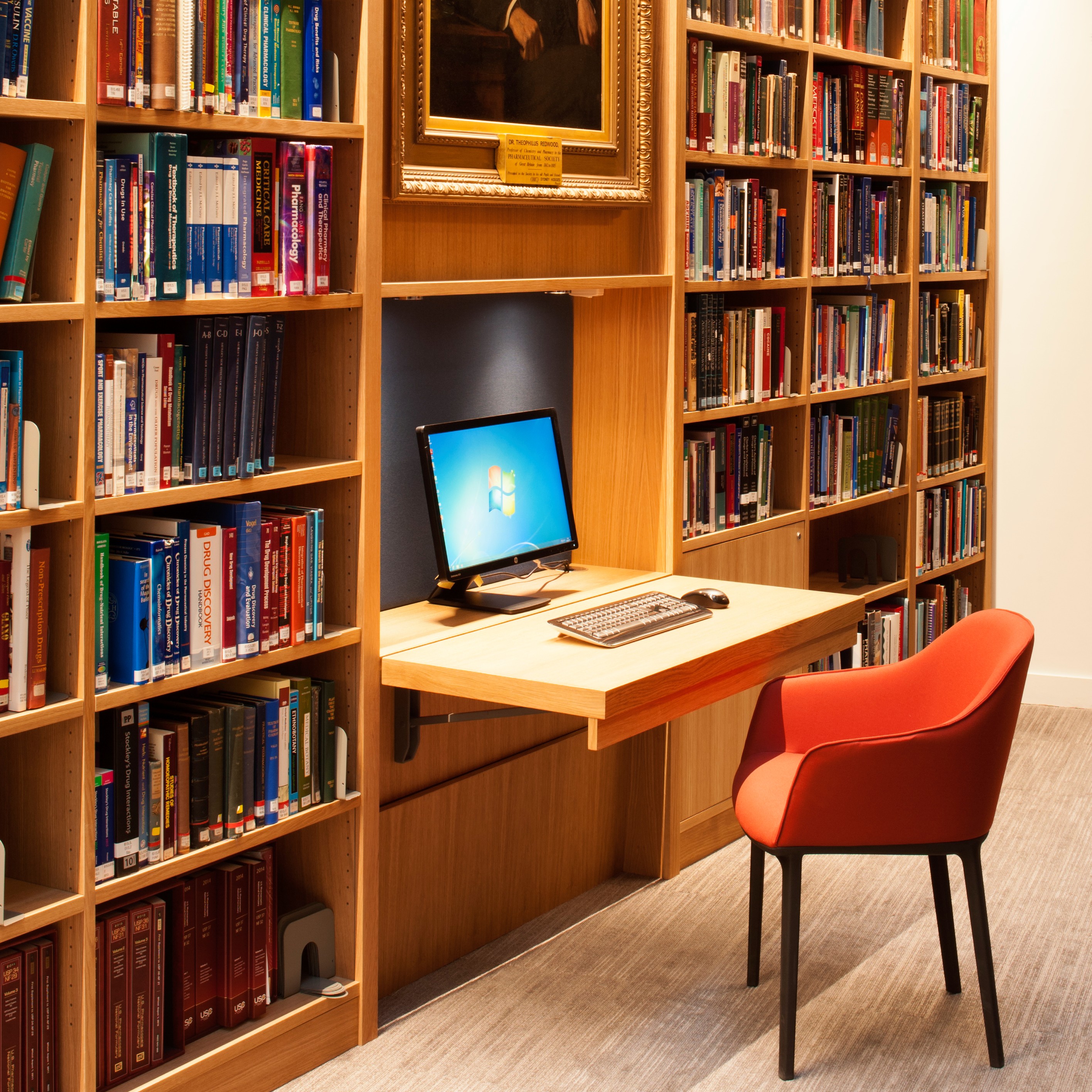 View of the Library showing a desk with a computer and chair, under a framed portrait of Theophilus Redwood, surrounded by wooden shelves holding books.